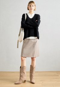 Suéter de punto negro, camisa de color crema con cuello, falda lápiz beige y botas de vaquero beige adornadas con un tacón cuadrado. El bolso es de color gris claro.