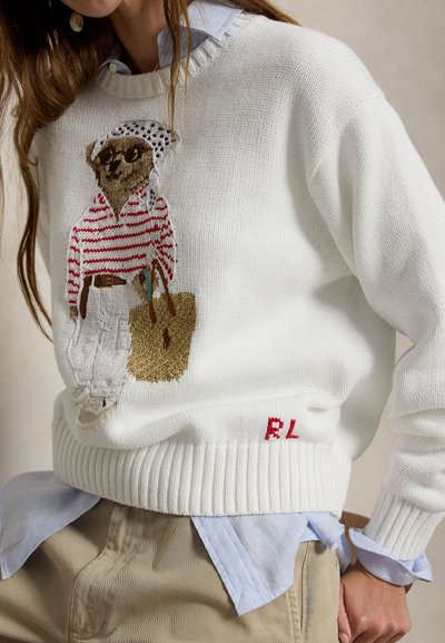 Λευκό πλεκτό πουλόβερ με κεντημένη αρκούδα που φορά γυαλιά ηλίου, κόκκινη ριγέ πουκάμισο, λευκό παντελόνι και κρατά υφαντή τσάντα tote, φορεμένο πάνω από μπλε πουκάμισο.