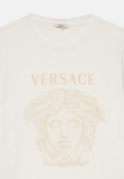 Λευκό t-shirt Versace με μπεζ γραφικό κεφαλής Μέδουσας και λογότυπο Versace πάνω από το σχέδιο στο κέντρο μπροστά.