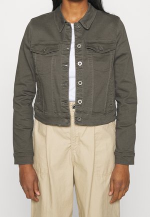 Woman wearing a dark olive buttoned jacket with flap pockets over a white top and beige high-waisted pants, standing against a plain background.