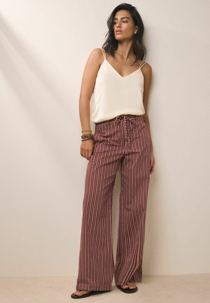 Žena oblečená v bielom tielku bez rukávov s výstrihom do V, hnedé široké pruhované nohavice s vysokým pásom, náramky a čierne sandále, stojaca pri béžovej stene.