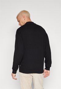 Suéter de punto negro con cuello redondo, mangas largas y puños acanalados. La tela parece suave con una textura agradable. Etiqueta de marca sutil en el dobladillo.