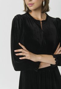Schwarzes Samtkleid mit hohem, gerafftem Ausschnitt, gesmoktem Taillenbund und Puffärmeln mit Rüschensaum. Mit Reißverschluss vorne.