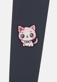 Leggings blu scuro con una toppa di un gatto in stile cartone animato rosa e bianco, con un fiocco, occhi espressivi e dettagli contornati. Tessuto dalla texture liscia.