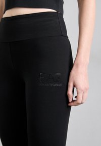 Colanți negri dintr-un material elastic cu talie înaltă, având o textură netedă și un logo din strasuri pe coapsă.