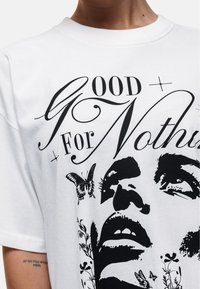Biała bawełniana koszulka z krótkim rękawem z czarnymi grafikami przedstawiającymi twarz, motyle oraz napis "Good For Nothing". Klasyczny dekolt i krótki rękaw.