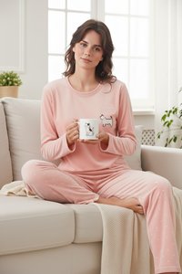 Kvinde i lyserødt nattøj med kattemotiv sidder med benene i kors på en beige sofa og holder et hvidt krus med en katteillustration.
