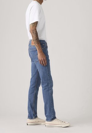 Person iført blå jeans, hvid t-shirt og hvide lave sneakers, der står sidelæns mod en ensfarvet baggrund.
