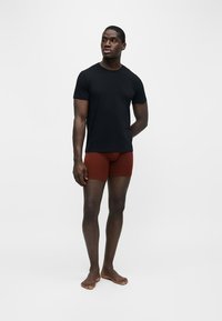 Homme debout pieds nus portant un t-shirt noir à manches courtes et un boxer couleur rouille sur un fond clair uni.