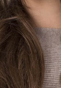 Les cheveux bruns ondulés tombent partiellement sur un pull gris clair à côtes avec une finition texturée. L'accent est mis sur les détails des cheveux et du tissu.