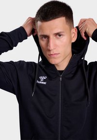 Zwart zip-up hoodie van zachte stof, met een voorkant rits, een capuchon met trekkoord en een logo op de linkerborst. Eenvoudig ontwerp en textuur.