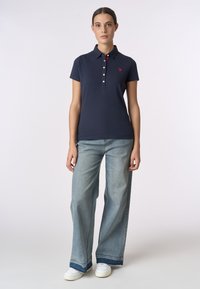 Polo navy con colletto e quattro bottoni, con un piccolo logo rosso. Abbinato a jeans ampi di colore azzurro chiaro e sneakers bianche.