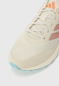 Lys beige sportssko med en tekstureret overdel, lyserøde accenter og en turkismønster. Har hvide snørebånd og tre orange striber på tungen.