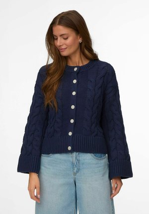 Kvinne iført en marineblå kabelstrikket knappet cardigan med lange ermer og lyseblå høytlivs jeans mot en hvit bakgrunn.