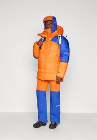 Chaqueta aislante naranja y azul con capucha, combinada con pantalones a juego. Presenta una textura acolchada, cremalleras y un diseño acolchado para mayor calidez.