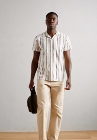 Chemise à manches courtes avec des rayures verticales en blanc, bleu, rouge et vert, associée à un pantalon beige clair. Le modèle tient un sac sombre.
