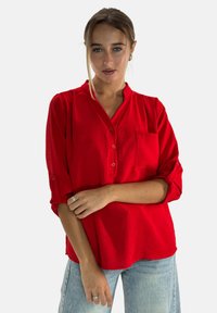Camicetta rossa con maniche arrotolabili, in tessuto testurizzato e con taschino frontale. Indossata con jeans azzurri chiari, che mostrano una vestibilità rilassata.