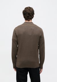 Hombre con cabello rizado castaño de espaldas, vistiendo un suéter de manga larga marrón y pantalones negros, contra un fondo liso claro.