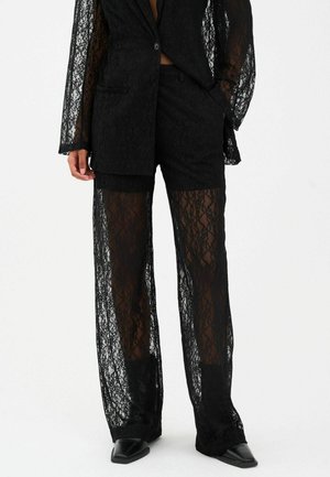Personne portant un blazer en dentelle noire et un pantalon large en dentelle transparente assorti, avec un short noir et des chaussures pointues en dessous.