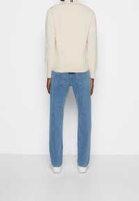 Persona che indossa jeans azzurri a gamba dritta, sneaker bianche e un maglione color crema, vista di spalle su uno sfondo bianco uniforme.