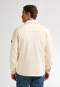 Camisa de manga larga color crema con cuello, botones laterales y un acabado texturizado sutil. Combinada con jeans azules, mostrando un estilo informal.