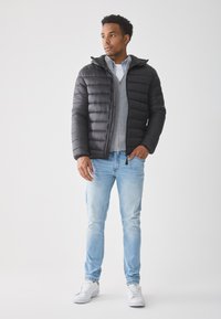 Hombre vestido con una chaqueta acolchada negra sobre un suéter gris, jeans azul claro y zapatillas blancas, de pie con una mano en el bolsillo, mirando hacia un lado.
