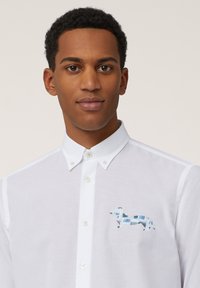 Chemise blanche à boutons avec une silhouette de chien brodée en bleu et en turquoise clair sur le côté gauche de la poitrine, fabriquée en tissu léger.