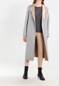 Grå ullkappa med en kontrasterande beige krage, lång rak design, två framknappar och bärs över en grå ribbad tröja och beige byxor.