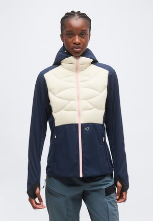 Mujer con chaqueta acolchada azul marino y crema con cremallera rosa y pantalones gris oscuro sobre un fondo claro liso.