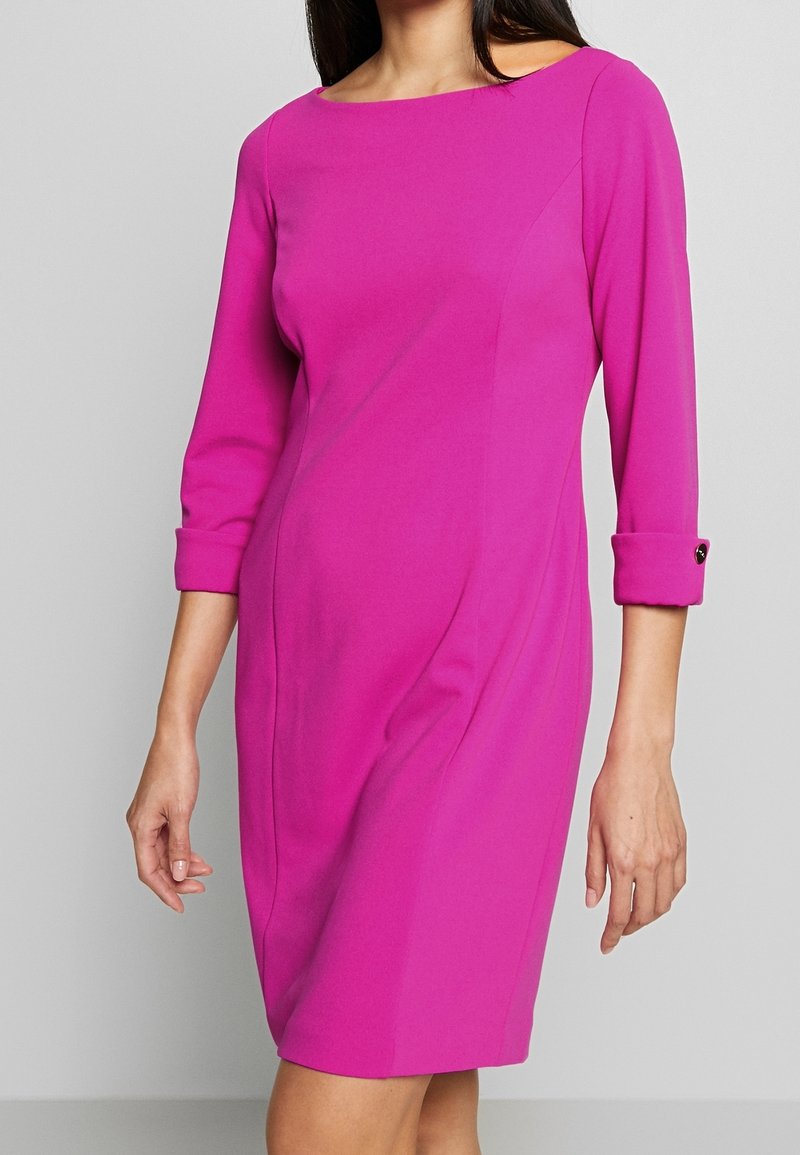 Femme portant une robe ajustée rose vif, longueur genou, manches trois quarts et poignets boutonnés, debout devant un fond gris uni.