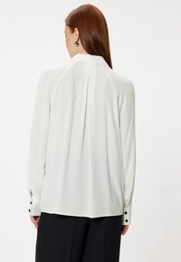 Weiße Bluse mit langen Ärmeln, die einen gefältelten Rücken und geknöpfte Manschetten aufweist. Hergestellt aus einem glatten Stoff mit lockerer Passform.