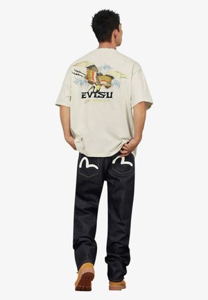 Uomo che indossa una camicia beige con motivo colorato di gru e jeans neri con cuciture bianche sulle tasche posteriori, in piedi con le mani nelle tasche.