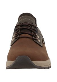 Bruna mockaskor med en mörk ovansida, kontrasterande sömmar, runda skosnören och en gummisula med texturerad detaljering. Logotyp på tungad.