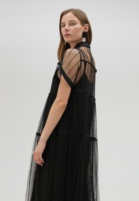 Robe noire transparente en tulle superposé, à manches courtes et col haut. Présente des rayures verticales et une silhouette fluide. Accessoirisée avec des boucles d'oreilles.