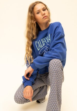 Blauwe trui met "CALIFORNIA" opdruk; grijze broek met patroon en diamantmotief. Gladde stof, ontspannen pasvorm en geribbelde manchetten.