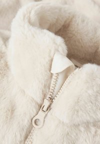 Pehme, koorevärvi kunstkarusnahast jakk kõrge kraega, nähtava tõmblukuga ja tekstuurse kangaga, mis annab pehme ja hubase välimuse.