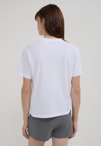 Donna con capelli castani che indossa una semplice maglietta bianca a maniche corte e pantaloni corto grigi, in piedi voltata di spalle contro uno sfondo grigio uniforme.