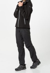 Sort softshelljakke med frontlynlås, parret med sorte bukser og sorte vandrestøvler. Outfitten har et slankt, funktionelt design.