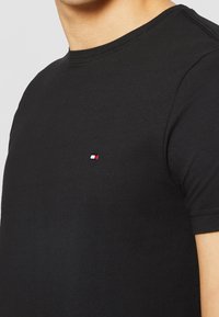Schwarzes Baumwoll-T-Shirt mit Rundhalsausschnitt und kurzen Ärmeln, mit einem kleinen roten, weißen und blauen Logo auf der linken Brustseite.