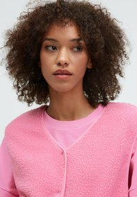 Rosa Fleece-Weste mit strukturierter Oberfläche, über einem einfarbigen rosa Sweatshirt getragen. Verfügt über einen runden Ausschnitt und einen einfachen Druckknopfverschluss.