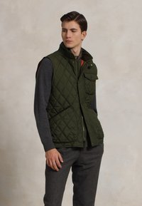 Pildīts olīvu zaļas krāsas veste ar augstu apkakli, priekšējo rāvējslēdzēju un pogu kabatām. Valkāta virs pelēka garām piedurknēm t-kreklu. Pielāgots piegriezums.