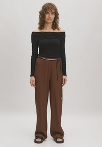 Zwarte off-shoulder top met geplooid detail, gecombineerd met losse, bruine textuur brede broek en zwarte sandalen. Voorkant gezien tegen een effen achtergrond.