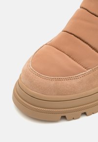 Zapatilla acolchada beige con parte superior de ante y textil suave, puntera redondeada y suela de goma texturizada con un patrón de tacos.