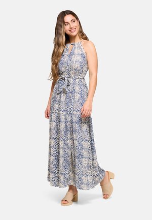 Mujer con cabello largo y ondulado que lleva un vestido maxi sin mangas con estampado azul y beige, y sandalias beige con tacón, sonriendo y mirando hacia un lado.