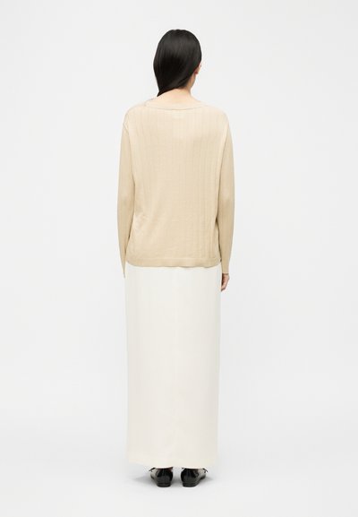 Femme aux cheveux longs et foncés, debout dos à la caméra, portant un pull côtelé beige, une longue jupe blanche et des chaussures noires sur un fond blanc uni.