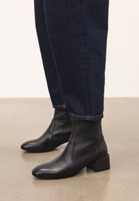 Mustad mustad nahast pahkluu saapad plokk-kandadega, kantud tumesiniste teksadega siledal beežil põrandal.