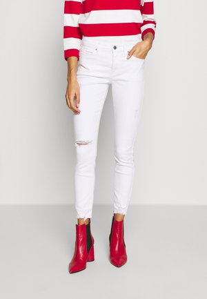 Witte skinny jeans met verwoeste kniezakken, gecombineerd met rode haklaarzen tot de enkel. De stof is denim met een gladde, licht rekbare textuur.