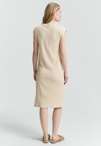 Abito beige, lungo fino al ginocchio, con una texture rugosa e maniche a cappuccio. Presenta un design semplice e una silhouette dritta, abbinato a sandali marroni.