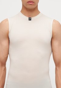 Chemise sans manches beige avec un col rond. Fabriquée dans un tissu texturé. Présente une petite étiquette noire au niveau du cou. Design ajusté.