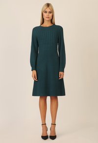 Teal strikket kjole med ribbet tekstur, der har en kabel-strikket overdel, lange ærmer og en flagrende nederdel, parret med sorte hælede sko.